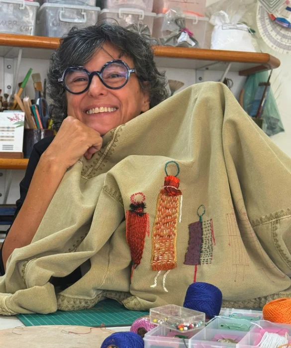 Lúcia Eid sorrindo ao lado de uma peça de tecido bordada à mão